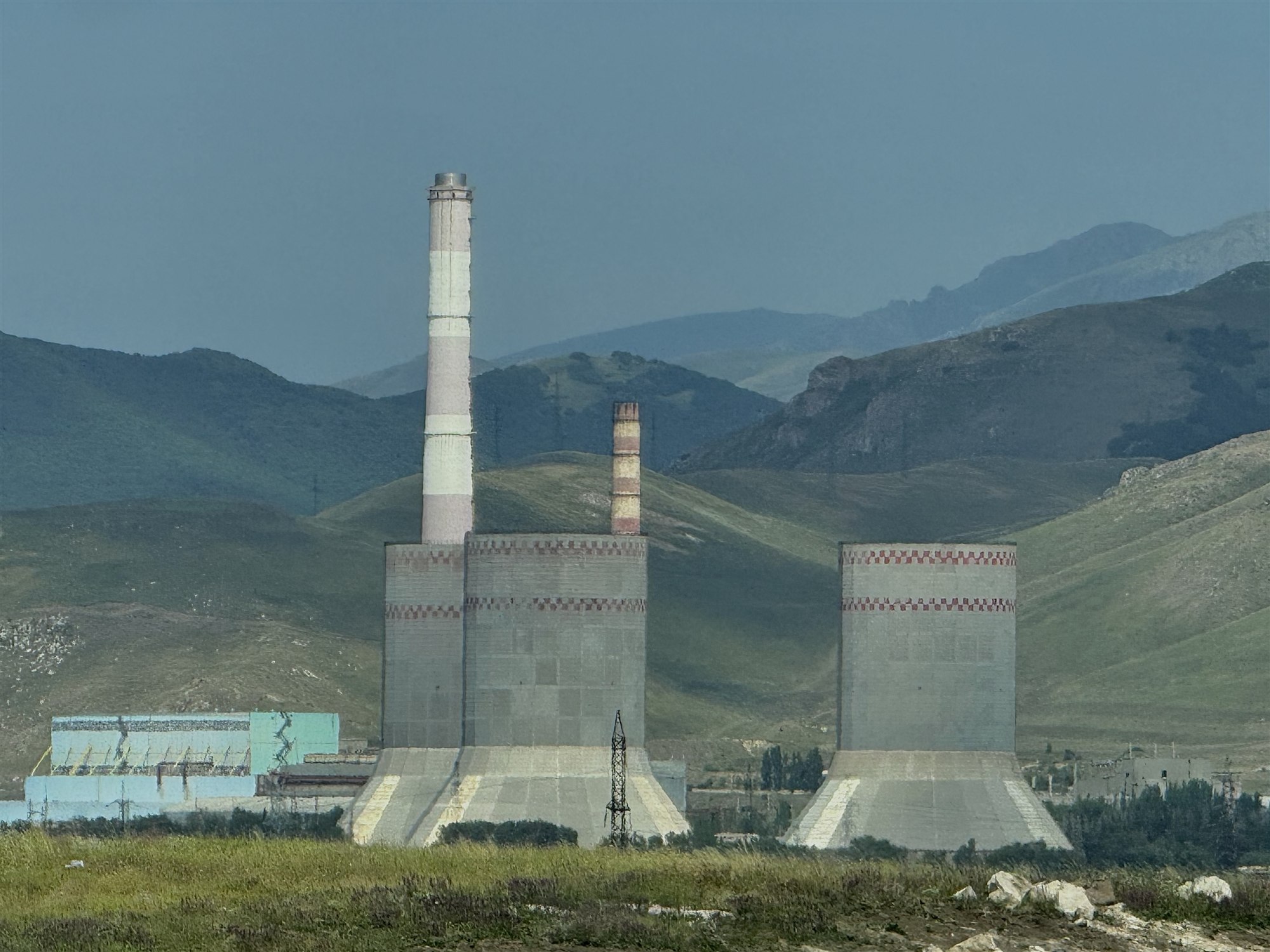 Two large concrete cooling towers and two banded chimneys of a thermal power plant, with green hills and mountains behind.